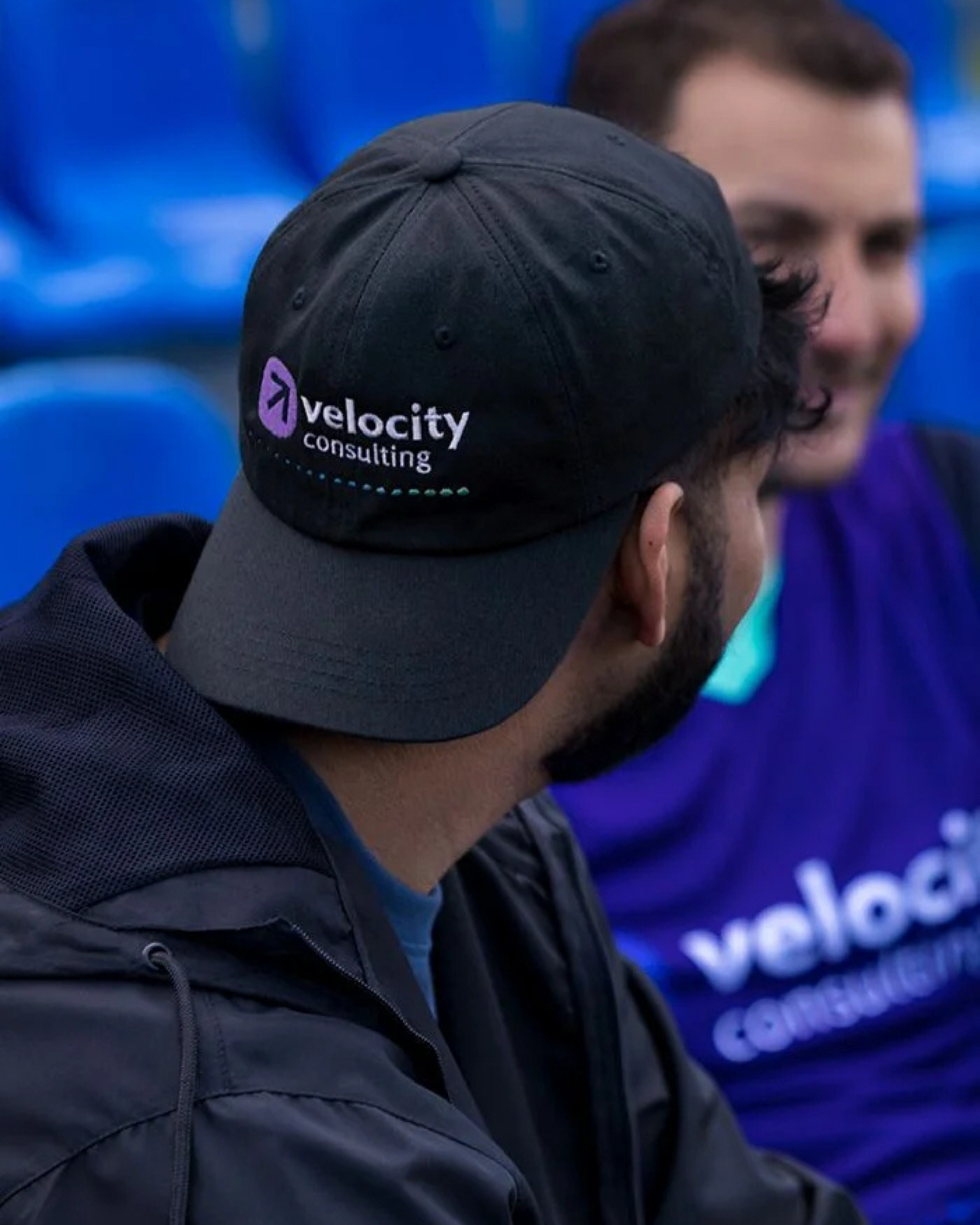 Person wearing a black cap with 'velocity consulting' logo, blurred background